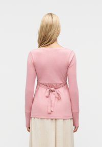 Γυναίκα με μακριά ξανθά μαλλιά που φοράει ροζ μπλούζα με μακριά μανίκια δεμένη με φιόγκο στο πίσω μέρος και μπεζ φούστα, στραμμένη προς τα πίσω σε μονόχρωμο φόντο.