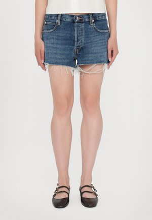 Slidte mørkeblå denimshorts båret med sorte flade sko med dobbelt strop på en person, der står mod en hvid baggrund.