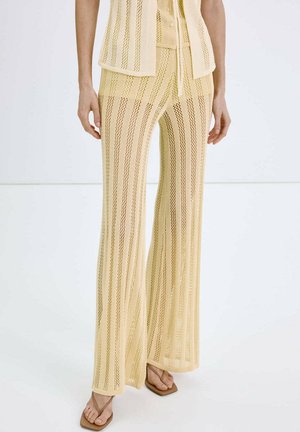 Modèle portant un pantalon en maille résille beige à rayures avec un haut sans manches assorti et des sandales à bout carré sur fond blanc.