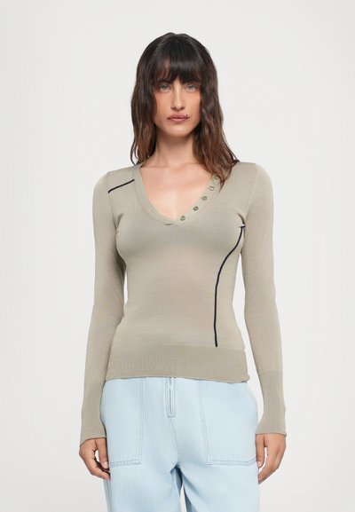 Beige Pullover mit langen Ärmeln und V-Ausschnitt, ausgestattet mit dunklen Akzentlinien und grünen Knöpfen entlang des Ausschnitts. Weicher Stoff, taillierte Passform.