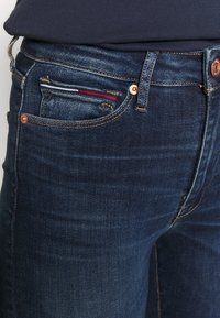 Jeans de mezclilla azul oscuro con un diseño clásico de cinco bolsillos, metalizado en cobre y un acento decorativo a rayas en el bolsillo delantero.