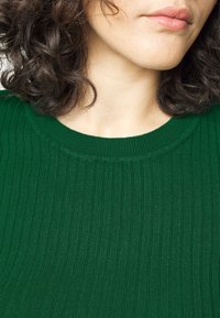 Maglione verde a coste con scollatura rotonda e texture morbida, parzialmente visibile su uno sfondo sfocato.