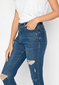 Jeans de mezclilla azul delgados con una cintura alta, que presentan desgarros en las rodillas, un leve desgaste y costuras en contraste.