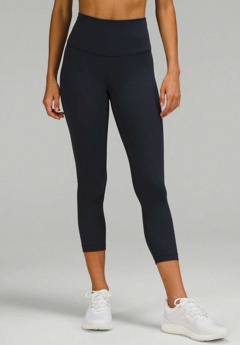 Högmidjade svarta leggings i stretchigt material, med en slät yta och kort längd, i kombination med vita sportskor.