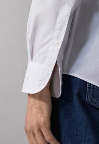 Close-up of a person’s arm wearing a white buttoned shirt cuff and blue denim jeans against a neutral background.