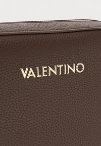Borsa in pelle marrone con superficie testurizzata e logo "Valentino" in oro. I bordi sono cuciti per resistenza e dettaglio visivo.