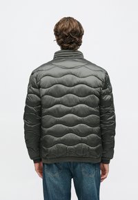 Μπουφάν με επένδυση σε σκούρο πράσινο χρώμα με κυματιστό σχέδιο με καπιτονέ, ψηλό γιακά, ελαστικές μανσέτες και τελείωμα. Το υλικό φαίνεται γυαλιστερό και ελαφρύ.