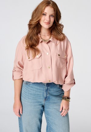 Vrouw met lang golvend rood haar, gekleed in een lichtroze overhemd met knopen en blauwe jeans, glimlachend met haar armen ontspannen langs haar zij.