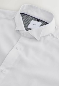 Biela košeľa s gombíkmi vyrobená z textúrovanej tkaniny. Má štandardný golier a tmavomodrý vzorovaný interiér pri výstrihu.