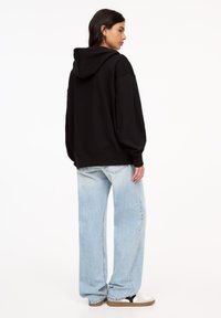 Hoodie noir à coupe décontractée, doté d'une capuche et de poignets côtelés, assorti à un jean large bleu clair et des baskets noir et blanc.