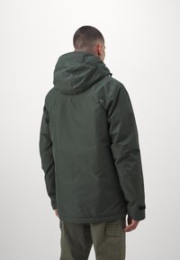 Donkergroene waterdichte jas met een capuchon, voorzien van een subtiele logo op de schouder en een verstelbare pasvorm. Geweven stof en naden zijn zichtbaar.