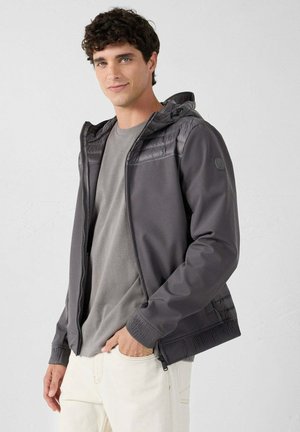 Grijze capuchonjas met gestructureerd bovenpaneel, rits aan de voorkant, geribbelde manchetten en zijzakken, gedragen over een effen grijs T-shirt.