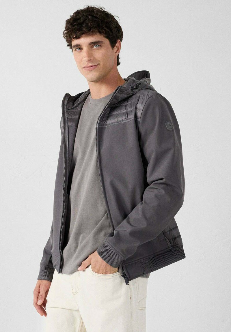 Grijze capuchonjas met gestructureerd bovenpaneel, rits aan de voorkant, geribbelde manchetten en zijzakken, gedragen over een effen grijs T-shirt.