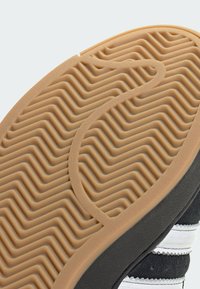 Gumená podrážka topánky s textúrovaným cikcak vzorom, béžovej farby a kontrastnými čiernymi okrajmi. Po boku sú biele pruhy.