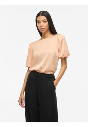 Femme portant un chemisier en satin pêche clair à manches bouffantes, rentré dans un pantalon noir large taille haute, posant avec une main dans la poche.