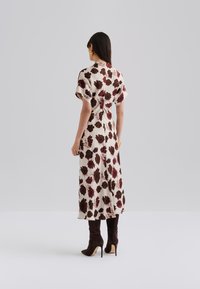 Maxime dress in a light beige fabric with a large red and dark brown floral pattern. Short sleeves and back closure, paired with brown heeled boots.