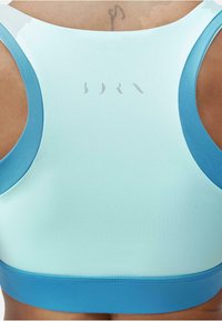 Sutien sport de culoare albastru deschis cu o textură netedă, având un tiv de culoare albastru mai închis și un branding subtil pe spate. Proiectat pentru îmbrăcăminte activă.