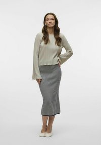 Lys beige, langermet plissert topp kombinert med en tettsittende, knelengd, marineblå og hvit stripet skjørt. Modell iført hvite leiligheter.