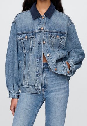 Femme portant une veste en jean bleu clair avec un col foncé et un jean taille haute assorti, une main dans la poche de la veste, boutons avant partiellement boutonnés.