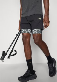 Μαύρες αθλητικές βερμούδες με σχέδιο στο τελείωμα, συνδυασμένες με γκρίζο μπλουζάκι και μαύρους ραχιαίους μπαστούνες. Μαύρα παπούτσια με υφασμάτινες σόλες.