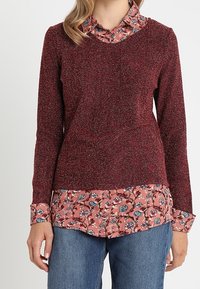 Maglione lavorato a maglia color bordeaux con un effetto testurizzato, indossato sopra una camicia rosa a stampa floreale, dotata di colletto e polsini con bottoni. Pantaloni in denim casual.