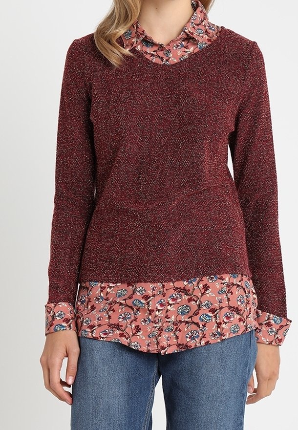 Maglione lavorato a maglia color bordeaux con un effetto testurizzato, indossato sopra una camicia rosa a stampa floreale, dotata di colletto e polsini con bottoni. Pantaloni in denim casual.