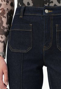 Jean en denim foncé à taille haute, avec des coutures jaunes et deux poches à l'arrière. Le tissu a une texture lisse et une coupe droite classique.