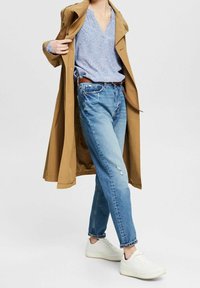 Impermeabile beige sopra una blusa blu a fantasia, jeans chiari strappati e sneakers bianche. L'outfit presenta una vestibilità rilassata e un design a scollo a V.