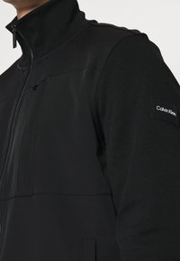 Musta vetoketjullinen takki, jossa on korkea kaulus, tyylikäs muotoilu, pehmeä kangas ja pieni Calvin Klein -logo hihassa.