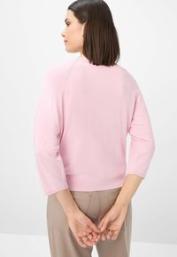Light pink, knitted, round-neck pullover with three-quarter sleeves and ribbed hem, worn with tan pants, hands clasped behind back.