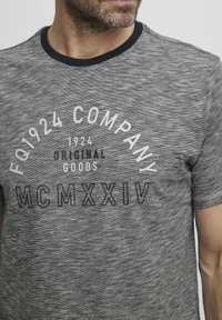 Sivka, kratka majica s kratkimi rokavi z teksturiranim vzorcem. Ima črn rebrast ovratnik in natisnjeno besedilo: "FQ1924 PODJETJE," "1924 IZVORNE BLAGOVNE ZNAMKE," "MCMXXIV."