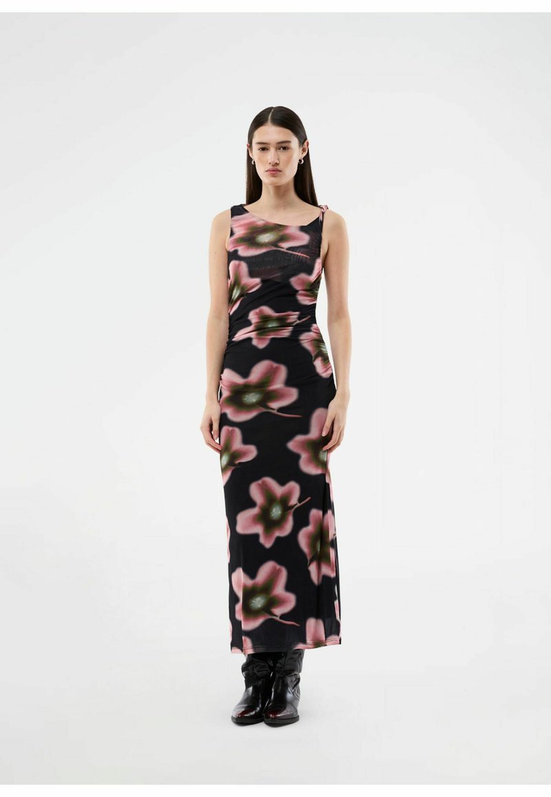 Femme debout, portant une robe maxi noire sans manches avec un grand imprimé floral rose et vert et des bottes noires, fond neutre.