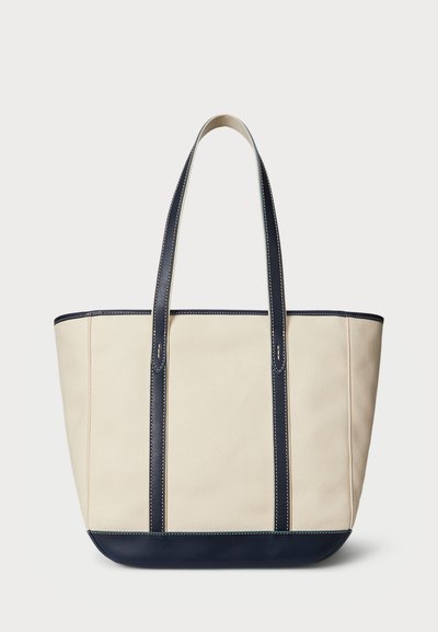 Plátená taška v krémovej farbe s námornícky modrými koženými akcentmi, ktorá má dva dlhé popruhy a viditeľné prešívanie pozdĺž okrajov.