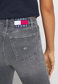Gray denim jeans with a branded patch featuring red, white, and blue colors. Includes two back pockets and subtle distressing.