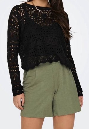 Femme portant un haut en crochet noir à manches longues et un short taille haute vert olive avec un collier chaîne, sur un fond uni.