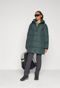 Chaqueta tipo abrigo larga y de color verde oscuro con capucha, que presenta detalles de costura. Combinada con pantalones oscuros y botas negras. Una mochila ligera está apoyada en el suelo.