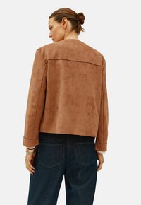 Giacca in suede marrone con vestibilità ampia, colletto largo e maniche lunghe. Presenta una spallina posteriore e dettagli con polsini a punta. Indossata sopra dei jeans scuri.