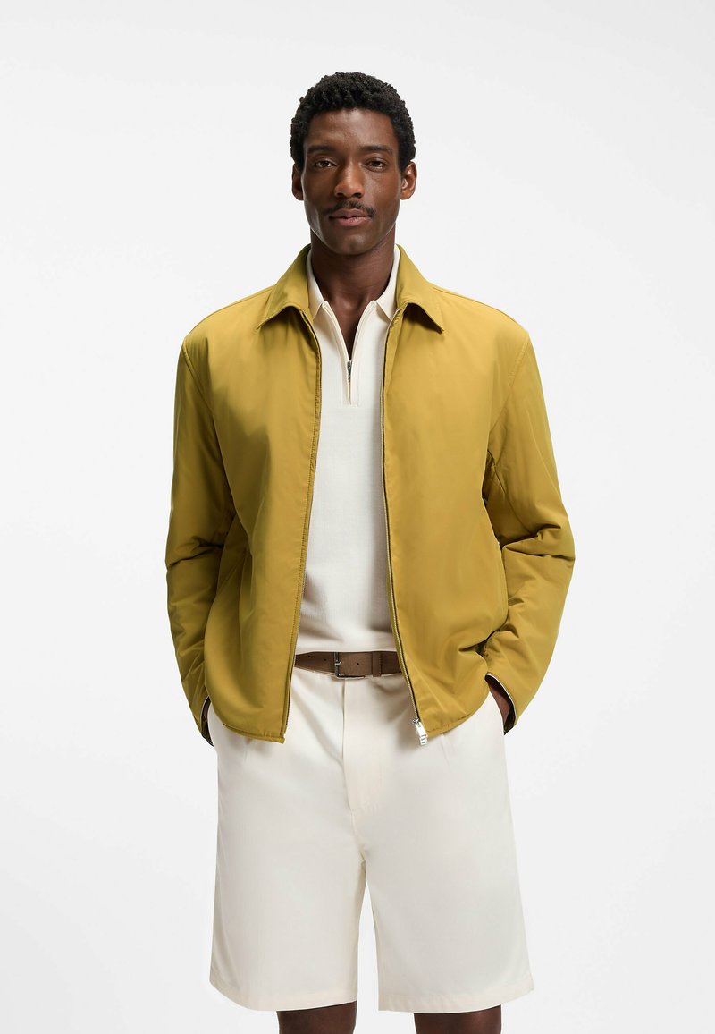 Homme portant une veste zippée jaune moutarde sur un polo blanc et un short beige, debout les mains dans les poches sur fond blanc.