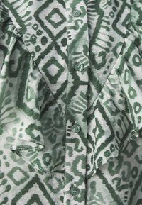 Tessuto verde a motivo geometrico con colletto a volant. Sei bottoni di un verde abbinato lungo la parte frontale.