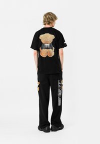 Camiseta de algodón negra con un gráfico de oso de peluche de gran tamaño en la parte posterior. Presenta pantalones holgados negros con parches de tela naranja y bolsillos.