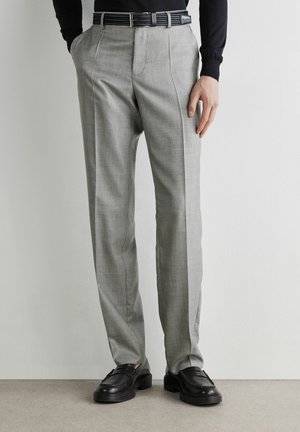 Uomo con pantaloni grigi chiaro su misura con pieghe frontali, cintura nera, maglia nera a maniche lunghe e mocassini neri in pelle che sta in piedi contro un muro bianco.