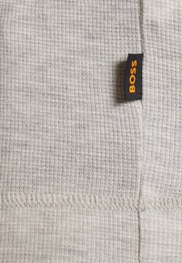 Tissu gris clair texturé avec une petite étiquette noire portant l’inscription « BOSS » en orange, cousue le long d’une couture.