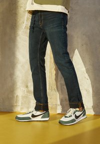 Sapatilhas Nike verdes e brancas com detalhes pretos, combinadas com jeans de ganga azul-escuro com costuras visíveis e bainhas encurtadas.