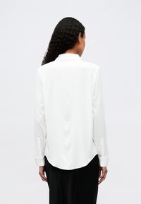 Chemise blanche à manches longues avec un col, texture lisse et ourlet incurvé ; accents de boutons visibles sur les poignets.