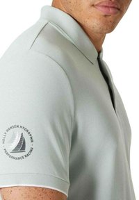 Polo shirt gris claro hecho de tela texturizada, con cuello y mangas cortas. Incluye un logo en la manga con un diseño de velero.