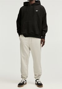 Schwarzer Kapuzenpullover mit Logo, Kängurutasche, kombiniert mit hellgrauen Jogginghosen und schwarzen Sneakers mit weißen Schnürsenkeln und Akzenten.