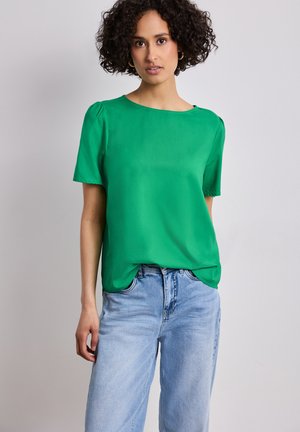 Femme aux cheveux bouclés portant des boucles d'oreilles anneaux dorés, un chemisier vert vif à manches courtes et un jean bleu clair sur un fond uni.