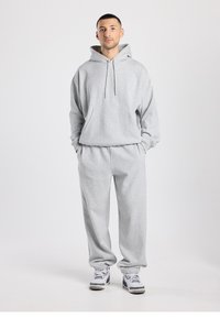 Sweat-shirt à capuche en coton gris avec une poche avant, associé à un pantalon de jogging gris assorti avec des ourlets élastiques. Le modèle se tient les mains dans les poches.
