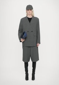 Blazer gris à double boutons, associé à un short assorti. Le mannequin porte une chemise noire, une casquette grise et des bottes noires montantes. Elle tient une pochette bleue.