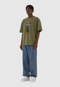 Oliven grøn oversized t-shirt med grafisk tryk af en ørn og teksten "Guardian Angel", parret med lyseblå denimjeans og hvide sneakers.